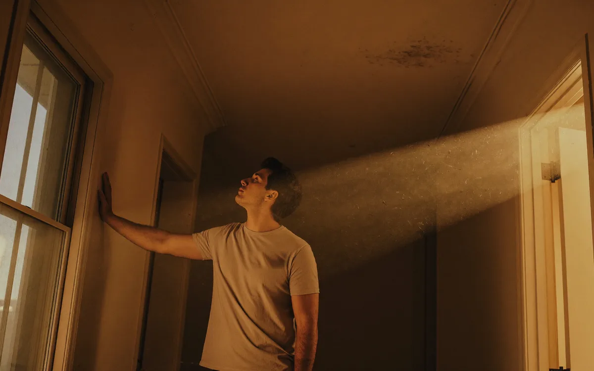 A man standing in the hallway of an older house with one hand on the wall, looking up toward the ceiling